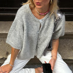 Femme portant un cardigan gris en tricot, collier rose, pantalon blanc, assise sur un trottoir. Mode décontractée, accessoires tendance.