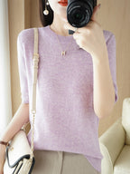 Femme en pull violet prend une photo avec un appareil photo, portant un collier doré et un sac à main beige. Mode féminine, accessoires élégants.