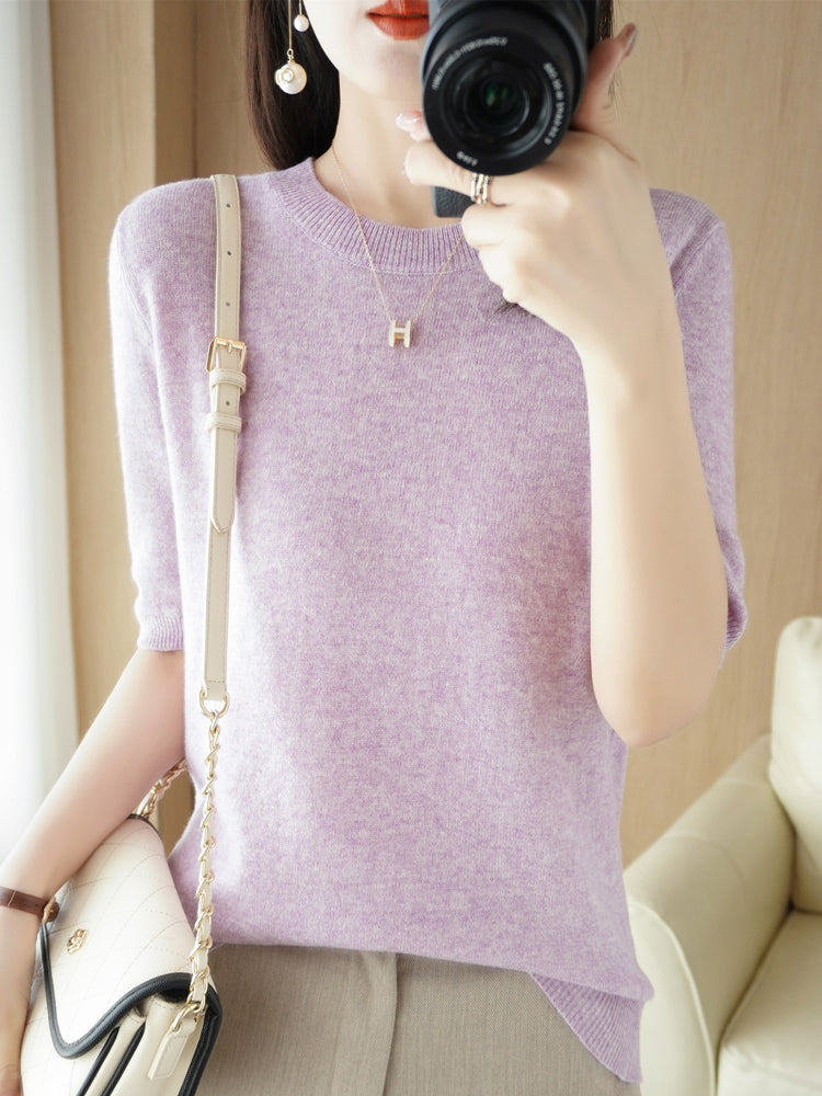 Femme en pull violet prend une photo avec un appareil photo, portant un collier doré et un sac à main beige. Mode féminine, accessoires élégants.