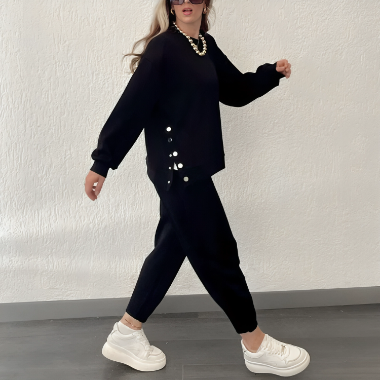 Femme en ensemble noir élégant avec boutons, collier doré, baskets blanches, marchant sur sol gris. Mode automne-hiver, style décontracté chic.