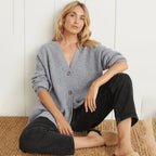 Femme assise portant un cardigan gris en maille avec boutons, pantalon noir et chaussons en fourrure, posant sur un tapis en jute. Mode décontractée.