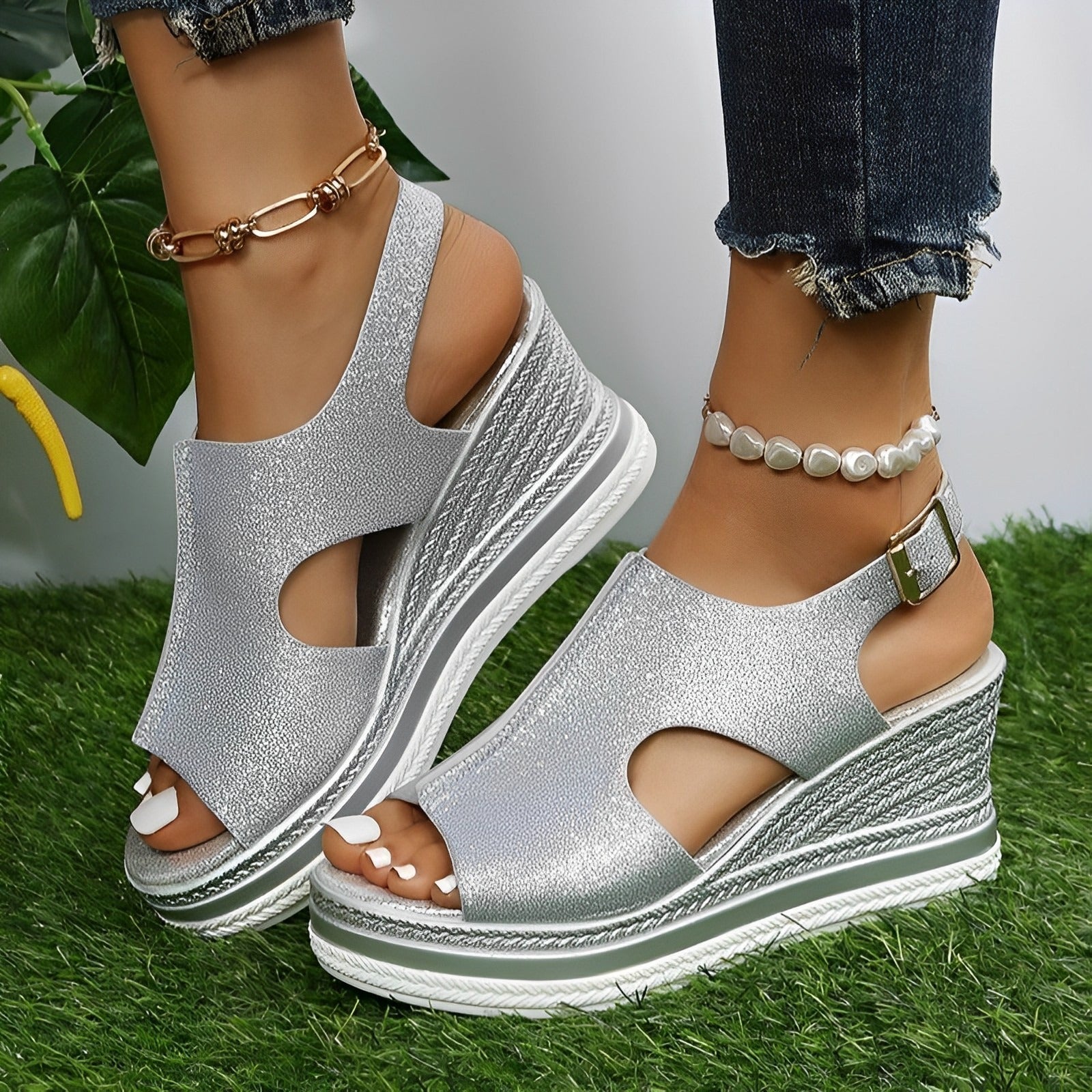Sandales compensées argentées pour femme avec talons plateforme, portées avec un jean et bracelets de cheville, posées sur gazon vert.