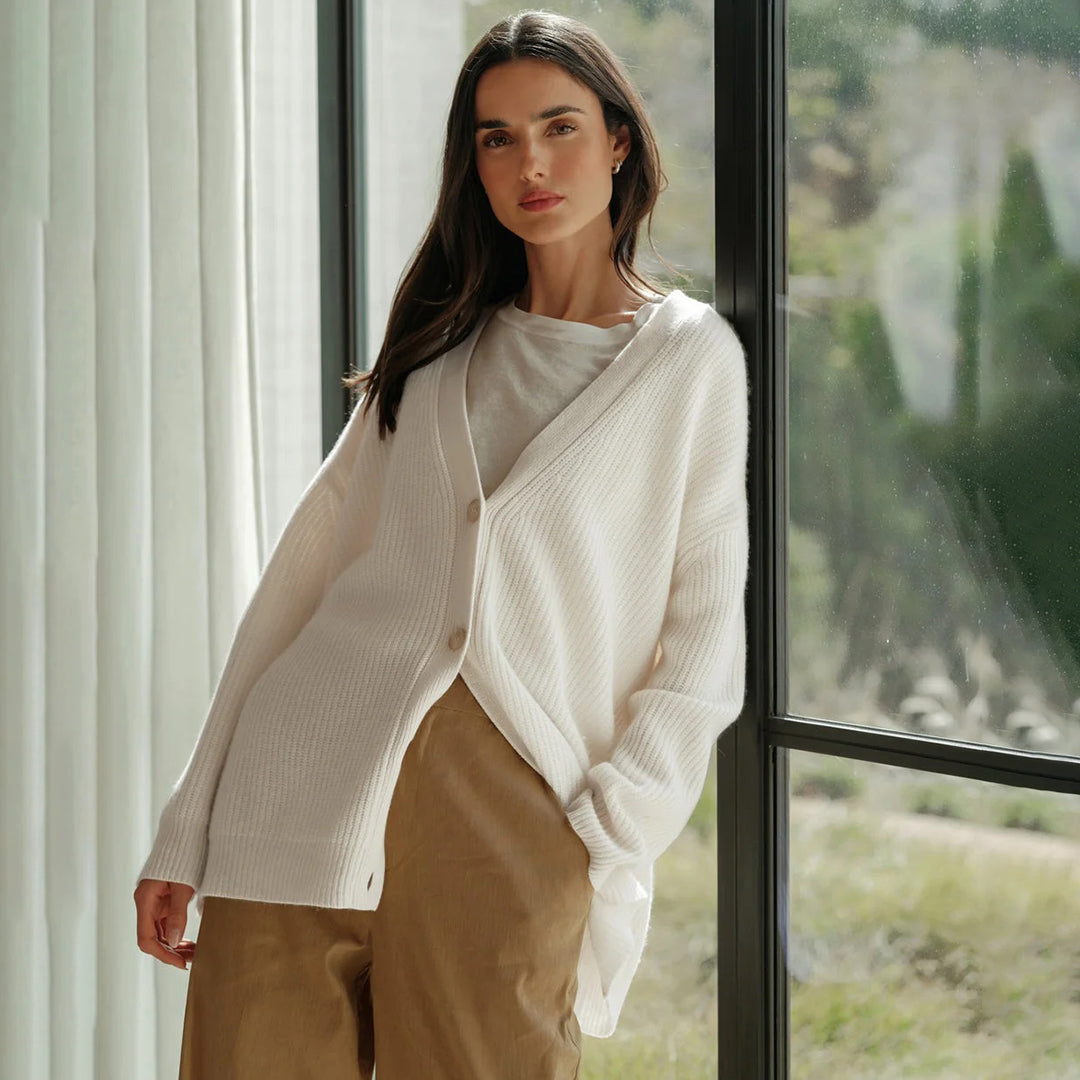 Femme portant un cardigan blanc en laine, debout près d'une fenêtre, lumière naturelle, style décontracté, mode automne-hiver, élégance simple.