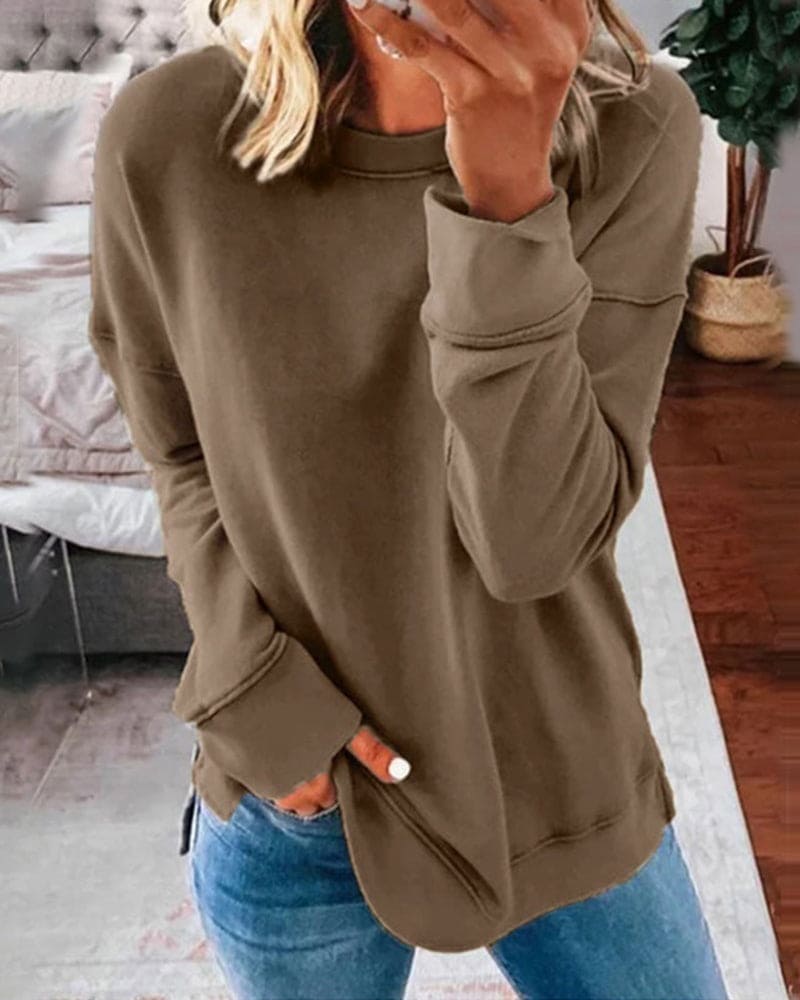 Femme en pull marron et jean bleu, tenant un smartphone devant un lit, plante en arrière-plan. Mode décontractée, intérieur moderne.