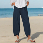 Personne portant un pantalon ample bleu marine et des sandales beiges sur une plage, ciel bleu en arrière-plan. Mode décontractée estivale.