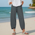 Personne portant un pantalon ample gris et des sandales sur une plage, avec l'océan en arrière-plan. Mode décontractée, tenue estivale, bord de mer.