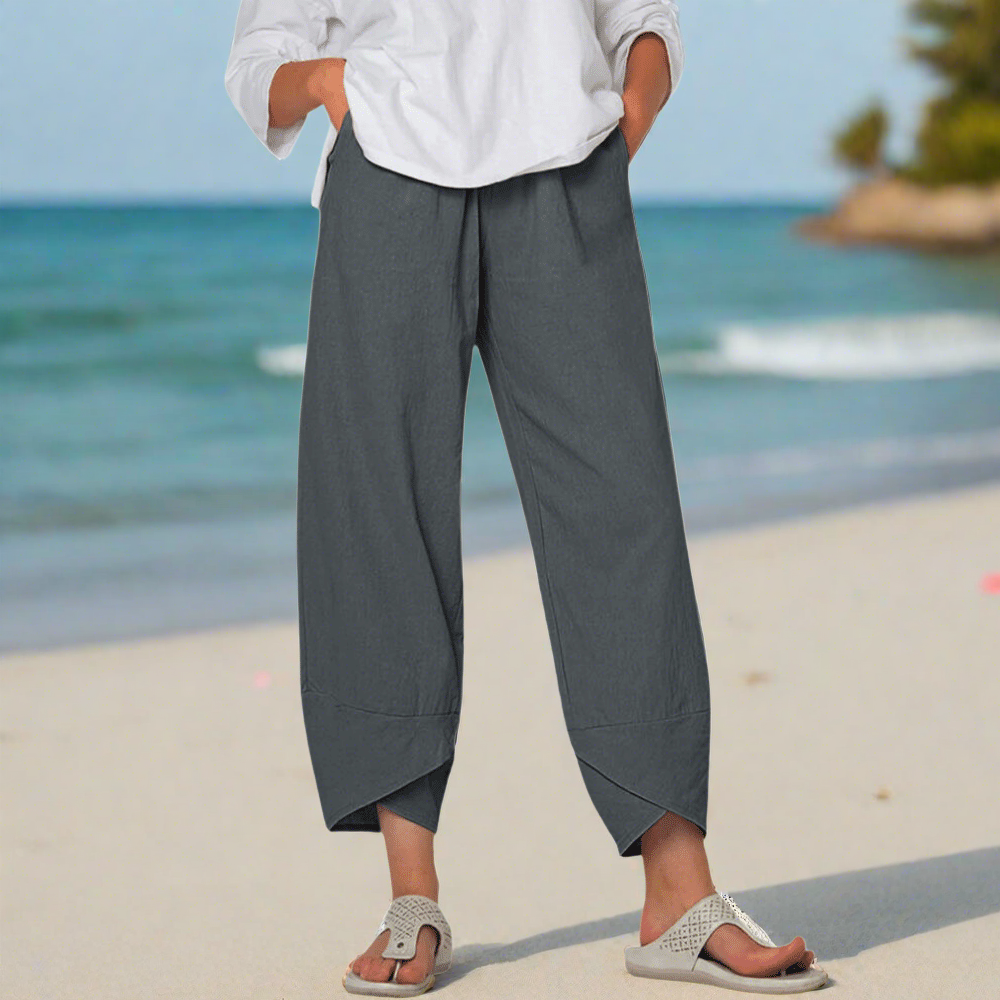 Personne portant un pantalon ample gris et des sandales sur une plage, avec l'océan en arrière-plan. Mode décontractée, tenue estivale, bord de mer.