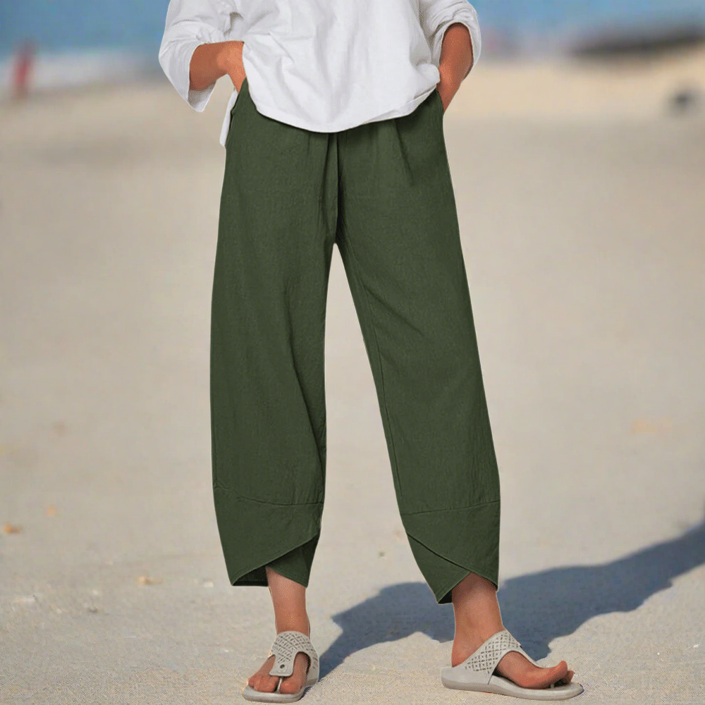Personne portant un pantalon vert ample et un haut blanc sur une plage, pieds dans des sandales beiges. Mode décontractée, tenue estivale, style plage.