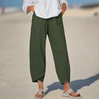 Personne portant un pantalon vert ample et un haut blanc sur une plage, pieds dans des sandales beiges. Mode décontractée, tenue estivale, style plage.