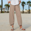 Pantalon beige ample en lin, porté avec une chemise blanche, sur une plage avec palmiers en arrière-plan. Mode estivale décontractée.