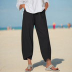 Personne portant un pantalon noir ample et des sandales beiges sur une plage ensoleillée, ciel bleu en arrière-plan. Mode décontractée estivale.