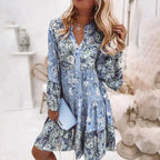 Robe fleurie bleue à manches longues, femme blonde tenant une pochette bleue, style bohème chic, intérieur lumineux, mode tendance.