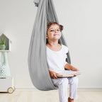 Enfant souriant assis dans un hamac gris suspendu, lisant un livre. Décor minimaliste avec jouet et plante en arrière-plan.