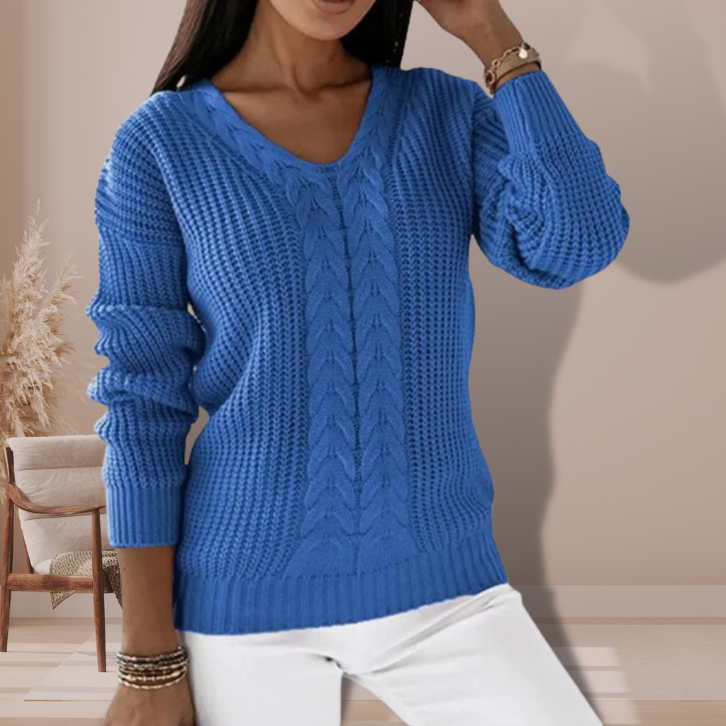 Femme portant un pull bleu en tricot torsadé, manches longues, col en V, posant dans un intérieur moderne avec chaise en bois et plante décorative.