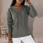 Femme portant un pull en tricot gris avec motif torsadé, manches longues, col en V, posant dans un intérieur moderne. Mode automne-hiver élégante.