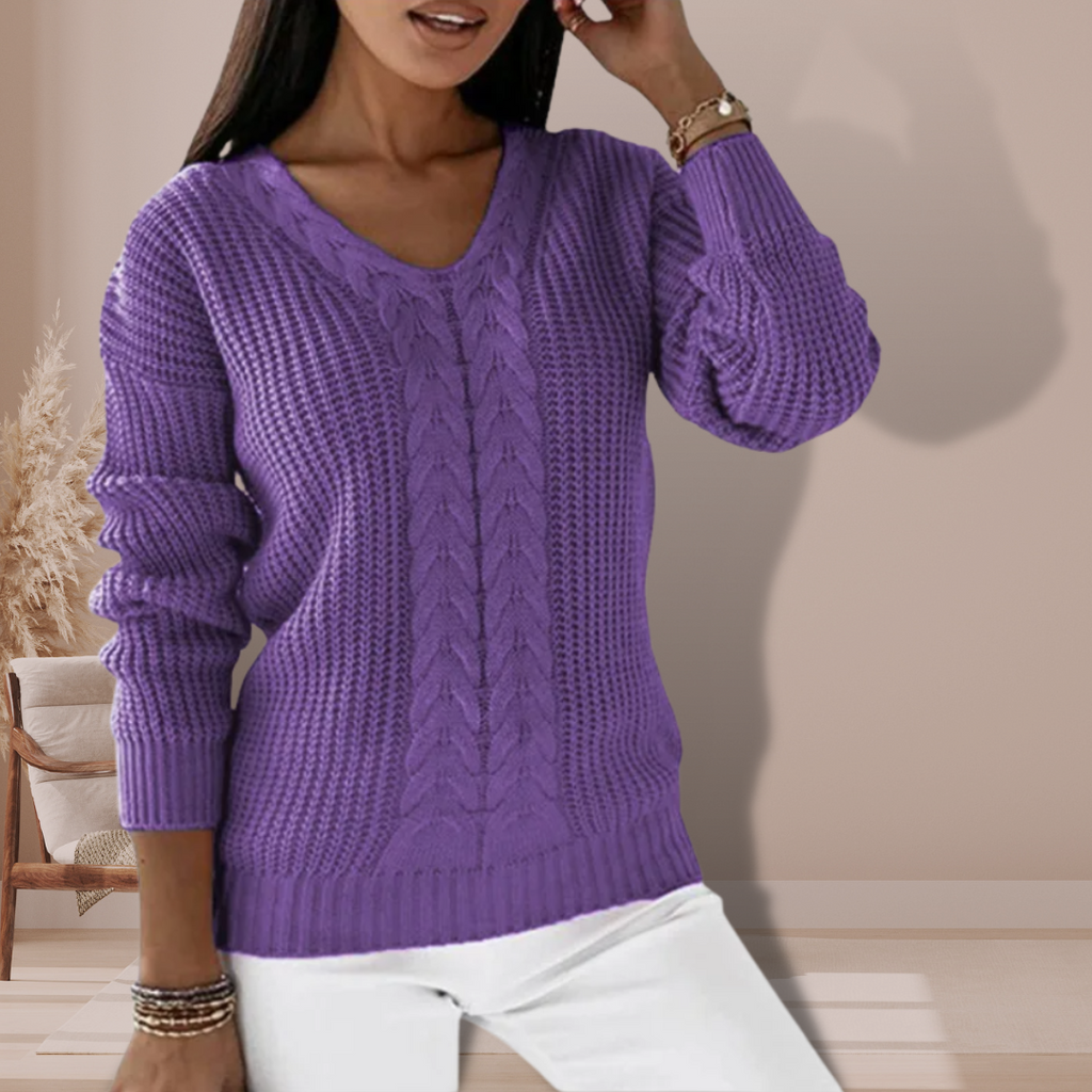 Femme portant un pull violet en tricot torsadé, manches longues, col en V, posant dans un intérieur moderne. Mode automne-hiver, style décontracté.