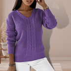 Femme portant un pull violet en tricot torsadé, manches longues, col en V, posant dans un intérieur moderne. Mode automne-hiver, style décontracté.