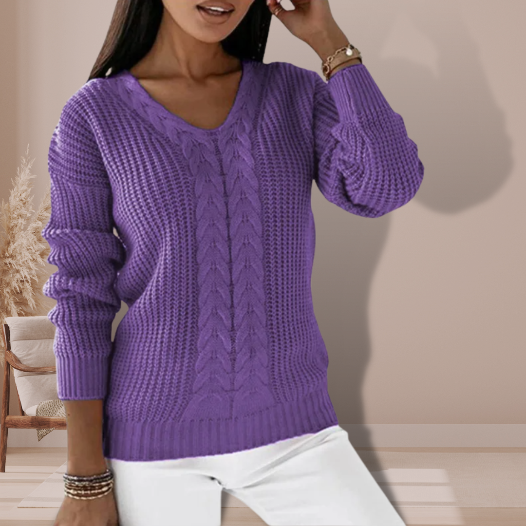 Femme portant un pull violet en tricot torsadé, manches longues, col en V, posant dans un intérieur moderne. Mode automne-hiver, style décontracté.