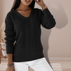 Femme portant un pull noir en tricot torsadé, manches longues, col en V, posant dans un intérieur moderne. Mode automne-hiver, style élégant.