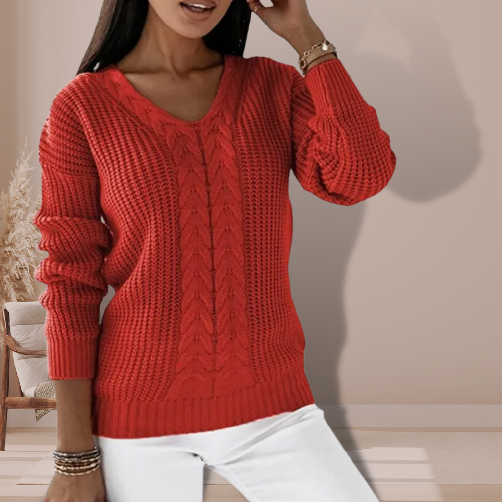Femme portant un pull rouge en tricot torsadé, manches longues, col en V, posant dans un intérieur moderne. Mode automne-hiver, style décontracté.