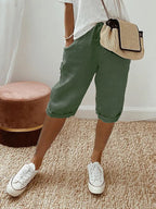 Femme portant un short vert en lin, t-shirt blanc, baskets blanches et sac à main en paille, posant sur un tapis beige. Mode décontractée estivale.
