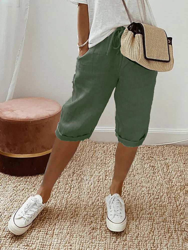Femme portant un short vert en lin, t-shirt blanc, baskets blanches et sac à main en paille, posant sur un tapis beige. Mode décontractée estivale.