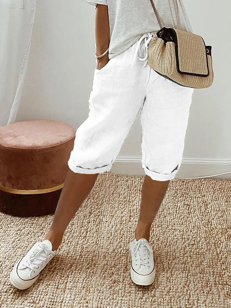 Femme en short blanc décontracté, t-shirt gris, baskets blanches, sac à main en paille, posant sur un tapis beige. Mode estivale chic et confortable.