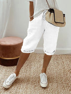 Femme portant un short blanc décontracté, t-shirt gris, baskets blanches et sac à main en paille, posant sur un tapis beige. Mode estivale chic.