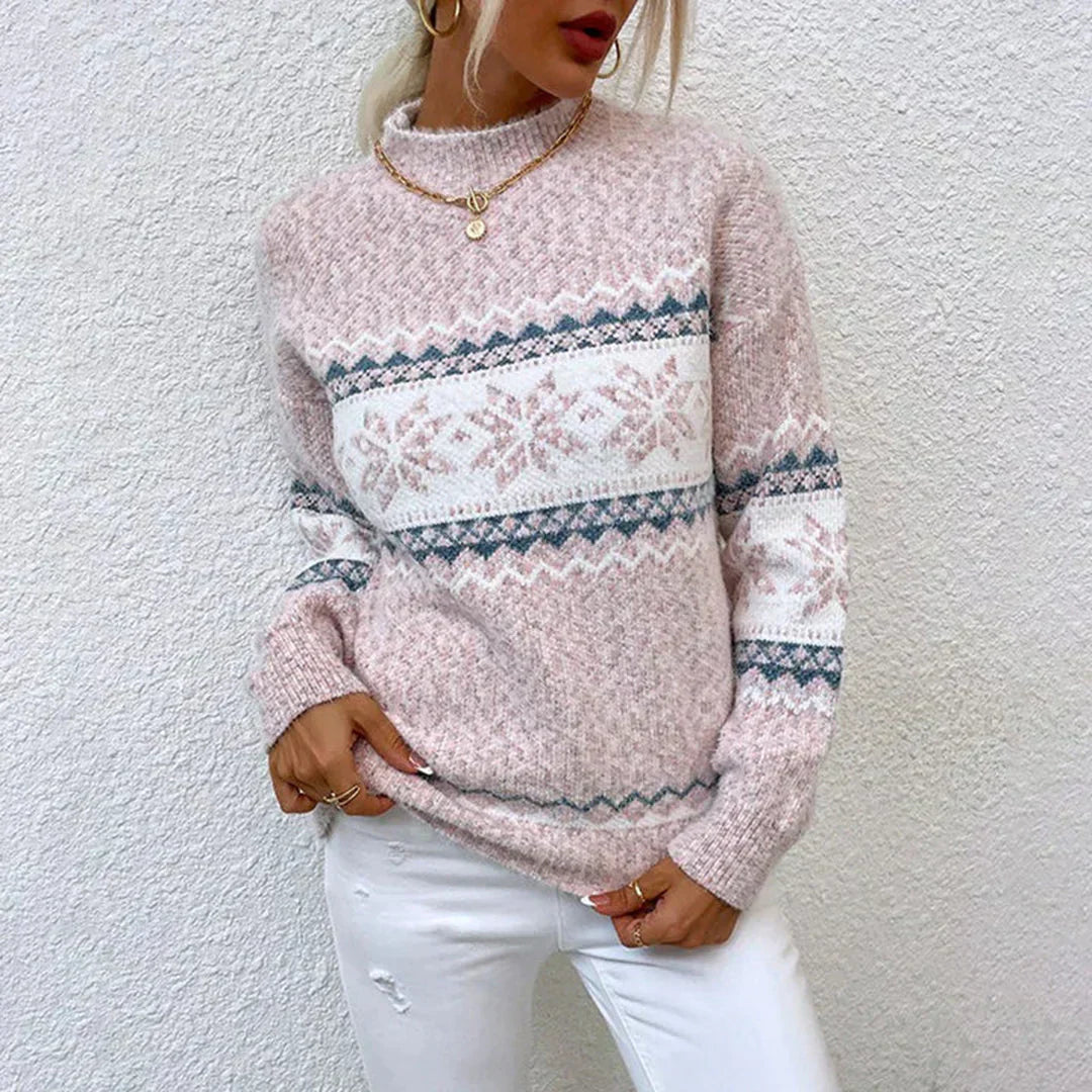 Femme portant un pull en tricot rose à motifs nordiques, col rond, manches longues, avec un collier doré, posant contre un mur blanc.