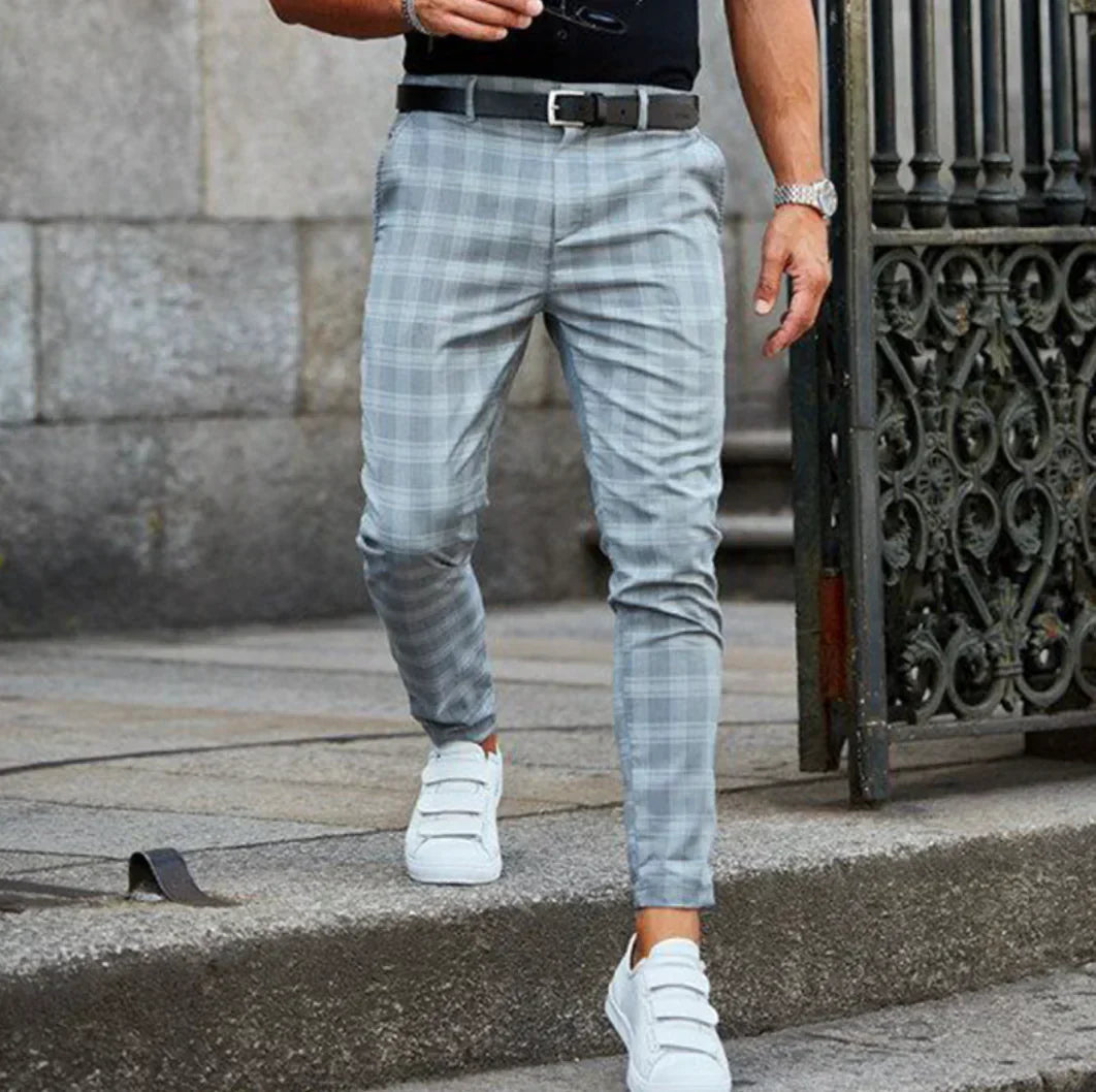 Homme portant un pantalon à carreaux gris, ceinture noire et baskets blanches, marchant sur un trottoir en pierre près d'une grille en fer forgé.