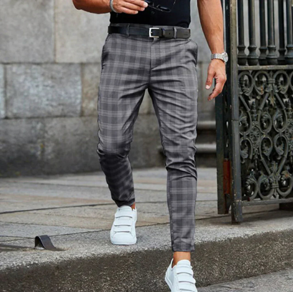 Homme portant un pantalon à carreaux gris et des baskets blanches, marchant sur un trottoir en pierre. Mode masculine élégante et décontractée.