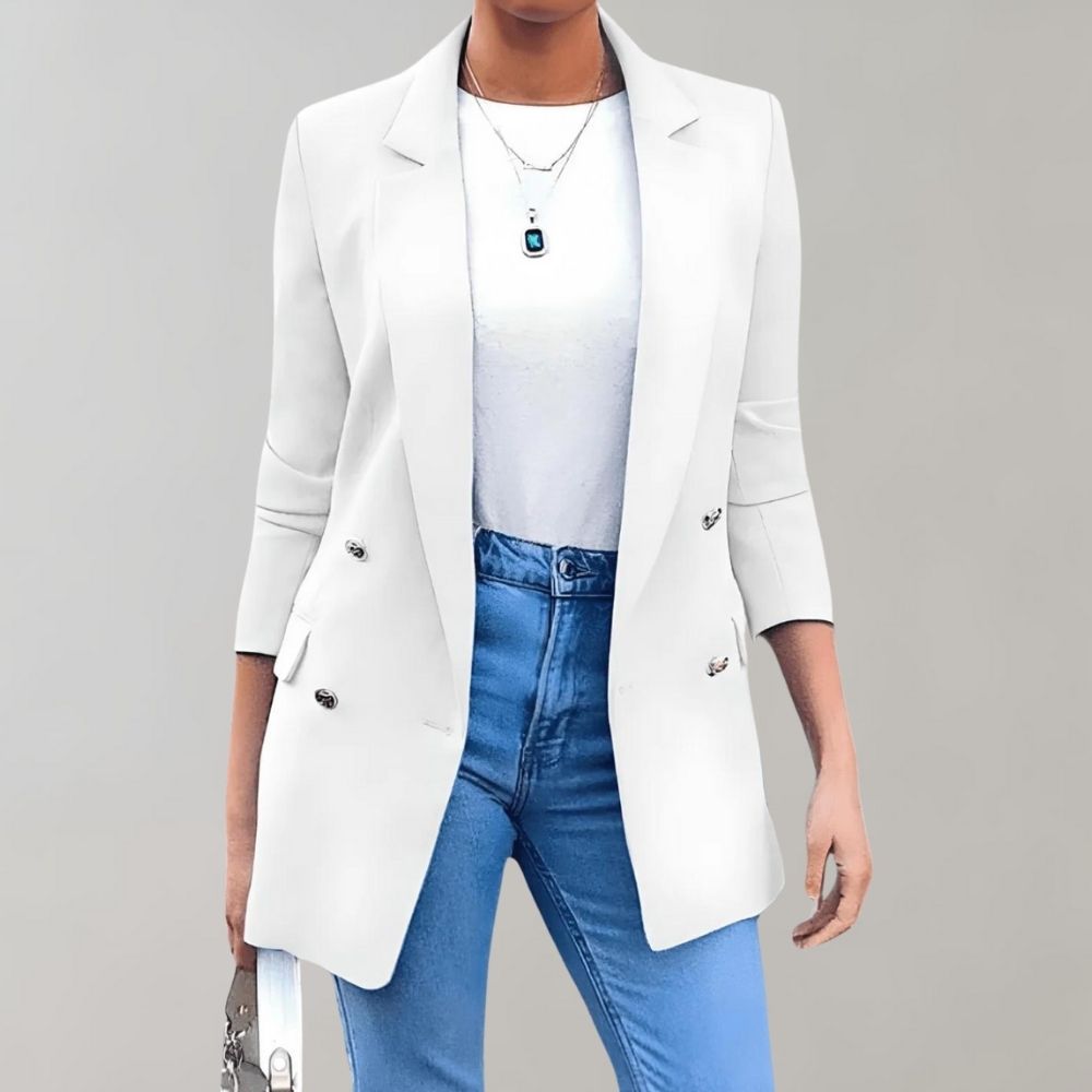 Femme portant un blazer blanc élégant, chemise blanche et jean bleu, accessoirisée d'un collier pendentif et d'un sac à main, sur fond gris.