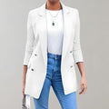 Femme portant un blazer blanc élégant, chemise blanche et jean bleu, accessoirisée d'un collier pendentif et d'un sac à main, sur fond gris.