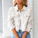 Femme portant une veste en jean blanche, manches retroussées, sur un t-shirt blanc, posant devant un mur en briques. Mode décontractée chic.