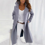 Femme portant un manteau gris élégant, chemise blanche et pantalon blanc, posant dans un intérieur moderne. Mode automne-hiver, style chic.