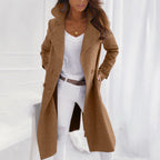Femme portant un manteau long marron, chemise blanche et pantalon blanc, posant dans un intérieur lumineux. Mode automne-hiver, élégance décontractée.