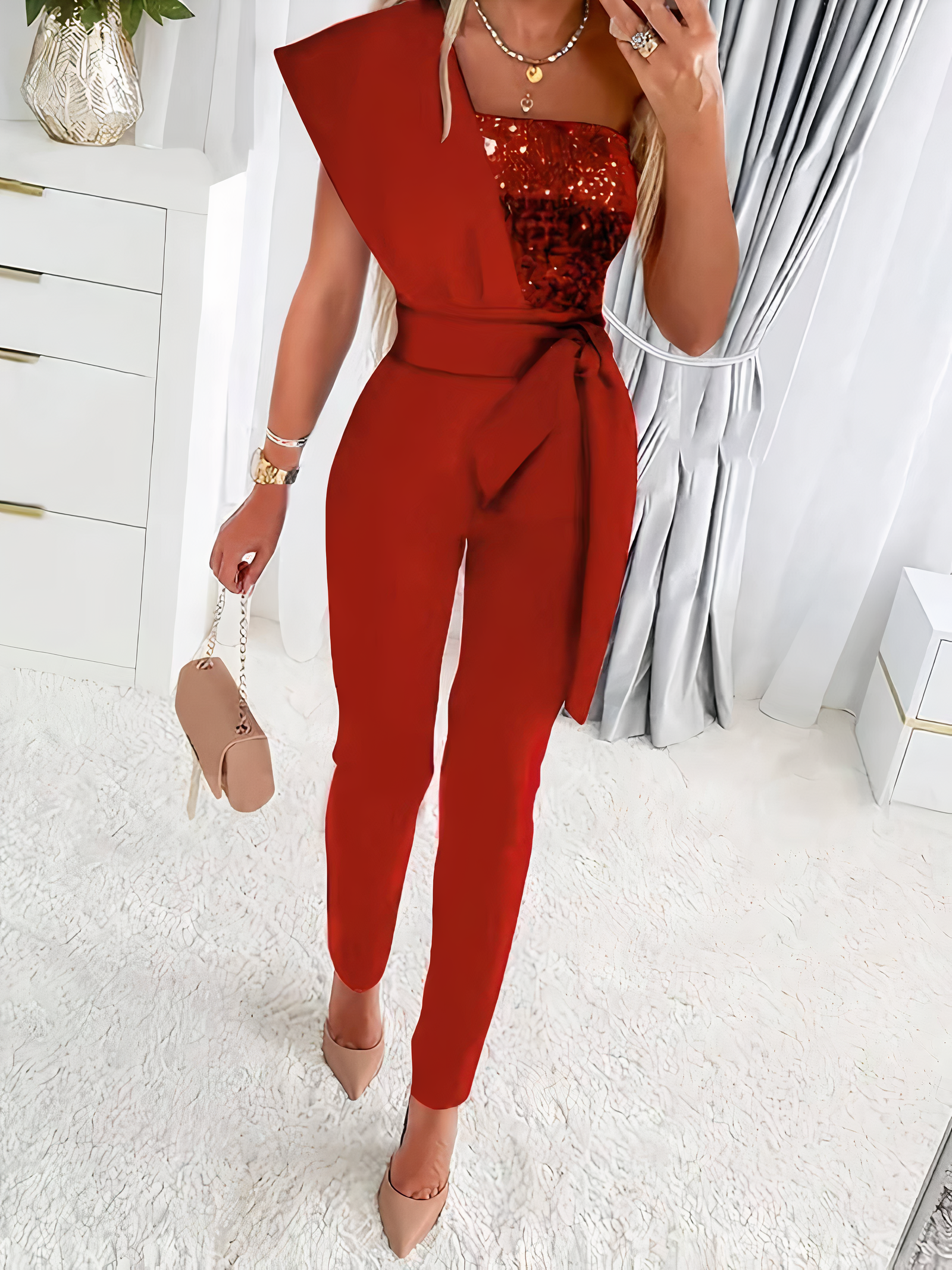 Combinaison rouge élégante avec paillettes, ceinture nouée, portée par une femme. Accessoires: sac à main beige, talons assortis. Mode chic et tendance.