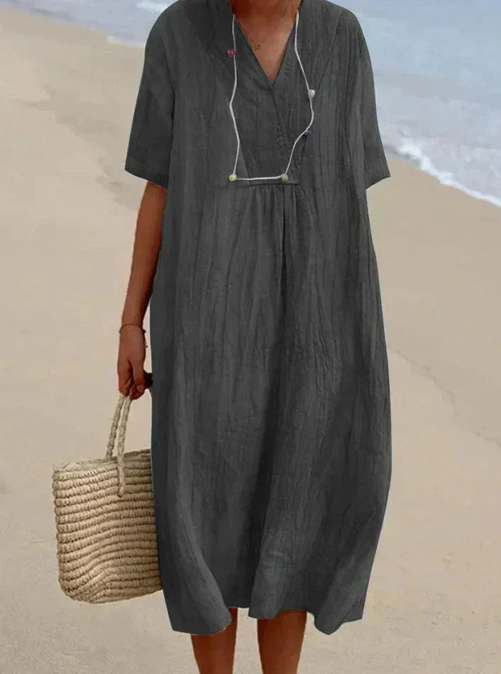 Robe d'été ample grise en lin, portée par une femme sur la plage, avec un sac en osier. Mode plage, tenue décontractée, style estival.