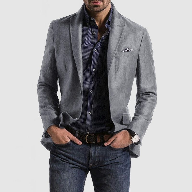 Homme portant une veste grise élégante, chemise bleue à motifs, jean décontracté, montre-bracelet et pochette de costume. Mode masculine chic.