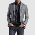 Homme portant une veste grise élégante, chemise bleue à motifs, jean décontracté, montre-bracelet et pochette de costume. Mode masculine chic.