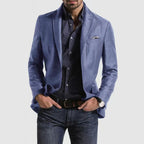 Homme portant une veste bleue élégante, chemise à motifs, jean décontracté, montre-bracelet et pochette, mode masculine moderne et chic.