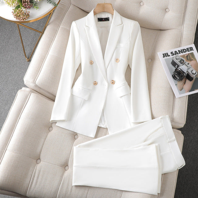 Costume blanc élégant pour femme, posé sur un canapé beige, avec une veste à double boutonnage et un pantalon assorti. Magazine de mode à côté.