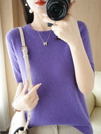Femme en pull violet en cachemire, tenant un appareil photo, portant un collier et un sac à main beige. Mode élégante, photographie lifestyle.