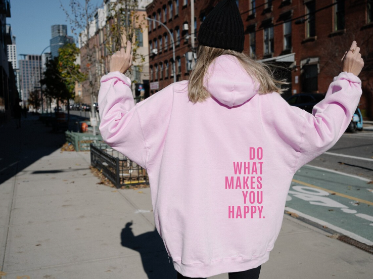Femme en sweat à capuche rose avec message "Do What Makes You Happy", marchant en ville, bâtiments en brique en arrière-plan, ambiance urbaine.