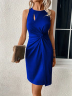 Robe bleue élégante sans manches avec nœud, femme tenant une pochette beige, devant une fenêtre. Mode féminine chic et moderne.
