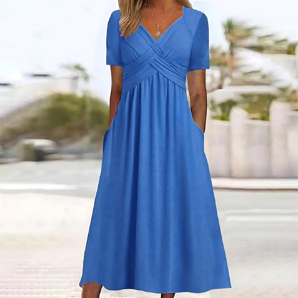 Robe longue bleue élégante avec manches courtes, col en V croisé, poches latérales, idéale pour l'été. Fond flou avec arbres et mur.