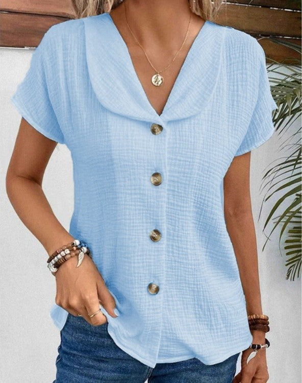Femme portant une chemise bleue à manches courtes avec col large et boutons en bois, accessoirisée de bracelets, devant un fond végétal.