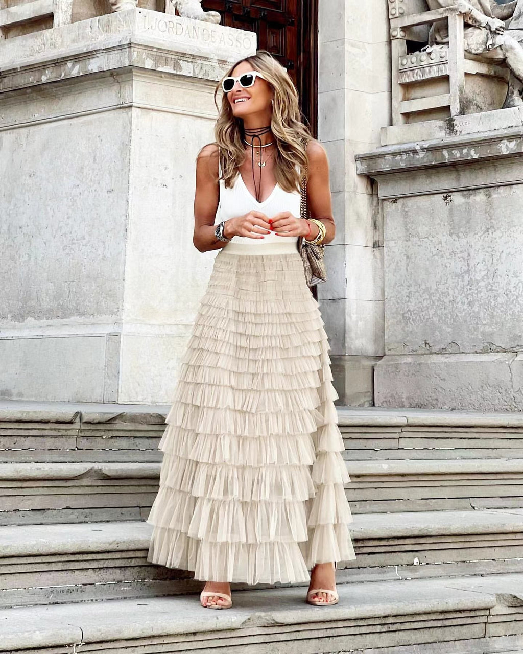 Femme élégante en jupe longue plissée beige et top blanc, posant sur des marches en pierre, lunettes de soleil blanches, style chic urbain.