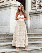 Femme élégante en jupe longue plissée beige et top blanc, posant sur des marches en pierre, lunettes de soleil blanches, style chic urbain.
