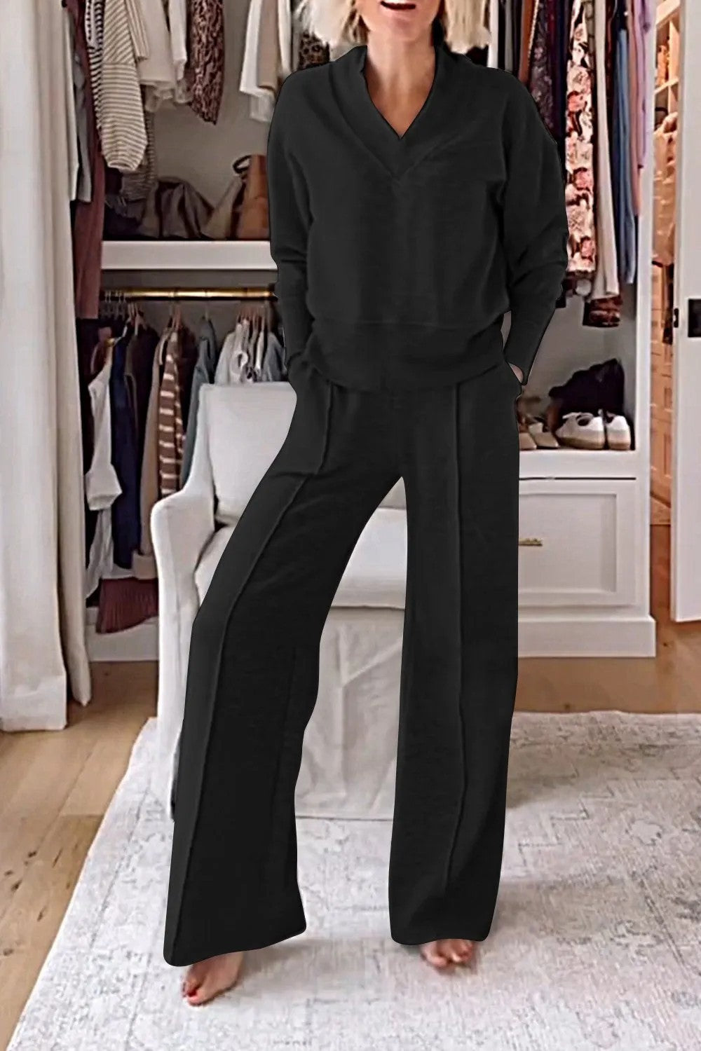 Femme en ensemble noir confortable, debout pieds nus dans un dressing moderne avec vêtements suspendus et étagères blanches en arrière-plan.